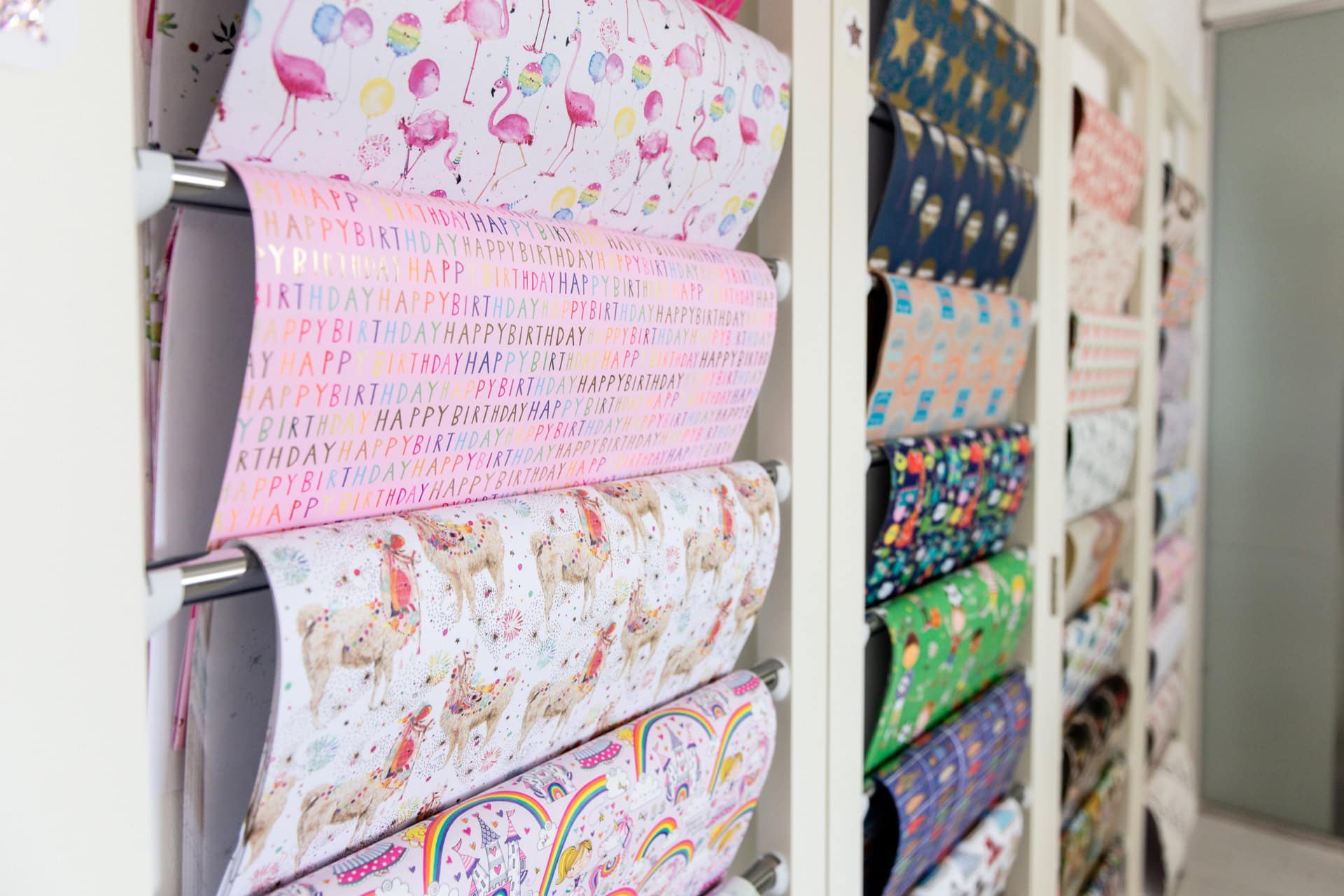 Vibrant gift wrap rolls featuring colorful patterns and festive designs displayed on a white rack at The Wraperie, a specialty wrapping paper store.