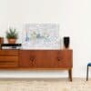 A sleek wooden sideboard featuring a musical stereo system with speakers, a decorative potted plant, and a colorful cityscape artwork, set against a minimalist white wall.