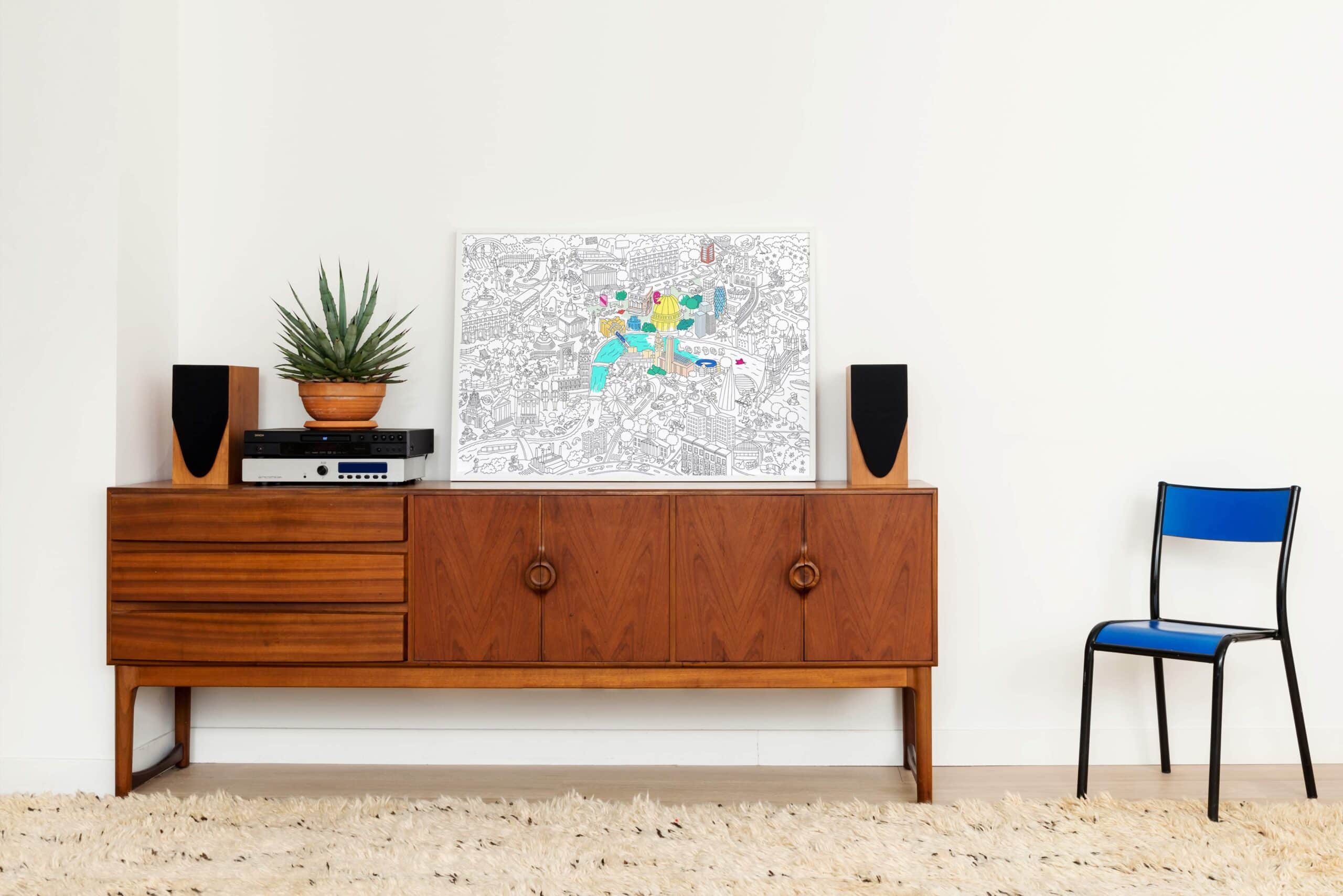 Modern interior decor with a vintage wooden sideboard, a potted succulent, a colorful city map artwork, black and wooden speakers, and a blue accent chair on a beige shag rug.