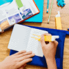 A close-up of a student's hand writing notes in a notebook on a wooden desk, surrounded by textbooks, highlighters, and stationery, emphasizing education and learning.