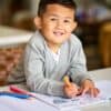 Child smiling while coloring at a table, kids activity, art and craft, happy kid, educational activity, creative childhood, learning through play, colorful markers, joyful learning environment.