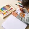 Colorful Crayons from The Wraperie on a white table with a child drawing and coloring a space-themed picture, showcasing fun art supplies and creativity.