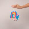 Colorful unicorn and rainbow character air freshener hanging from hand, fun and whimsical home decor, perfect for kids' rooms or playful spaces, from The Wraperie online store.