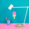 Pink popsicle happy face wall hanging hanging charm, desk lamp, colorful pens in blue container, fun colorful workspace decor, playful ice cream themed accessories, modern work from home setup, aesthetic office decor.