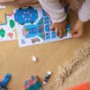 Colorful children's wooden house and city scene puzzle from The Wraperie with matching stickers and game pieces on wooden floor.