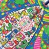 Bright colorful kids' beach mat featuring playful cartoon dinosaurs, umbrellas, and fun phrases like 