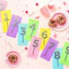 Brightly colored confetti cake toppers with glittery numbers 1-9 on a pink background, complemented by donuts, pink cups, and festive sprinkles, perfect for birthday celebrations and dessert decorating.
