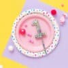 Bright pink paper plate with colorful sprinkles shaped as the number one, surrounded by confetti, candies, and balloons on pastel yellow and purple background for celebrating first birthday.