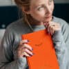 Relaxed woman holding bright orange wellness journal, smiling thoughtfully, indoors, casual gray sweatshirt, positive mindset, self-care, mental health, lifestyle, wellness, personal growth, The Wraperie.