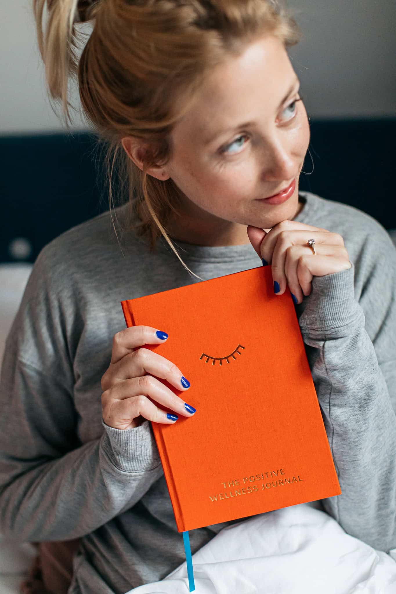 Relaxed woman holding bright orange wellness journal, smiling thoughtfully, indoors, casual gray sweatshirt, positive mindset, self-care, mental health, lifestyle, wellness, personal growth, The Wraperie.