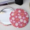 Bright pink floral fabric mirror compact with makeup brushes and jewelry on a white surface, ideal for beauty and cosmetic enthusiasts.