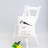 Handmade tote bag with bird illustration and inspiring quote, displayed on white distressed chair, complemented by colorful flowers, showcasing stylish and eco-friendly accessories at The Wraperie.