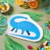 Colorful Dino Snack Plate featuring a blue dinosaur shaped paper plate, surrounded by tropical leaves, fresh fruit slices, and vibrant candies, perfect for kids' birthday parties and dinosaur-themed events.