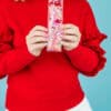 Bright woman in red sweater holding colorful glitter-filled beverage container with pink straw, smiling against a blue background.