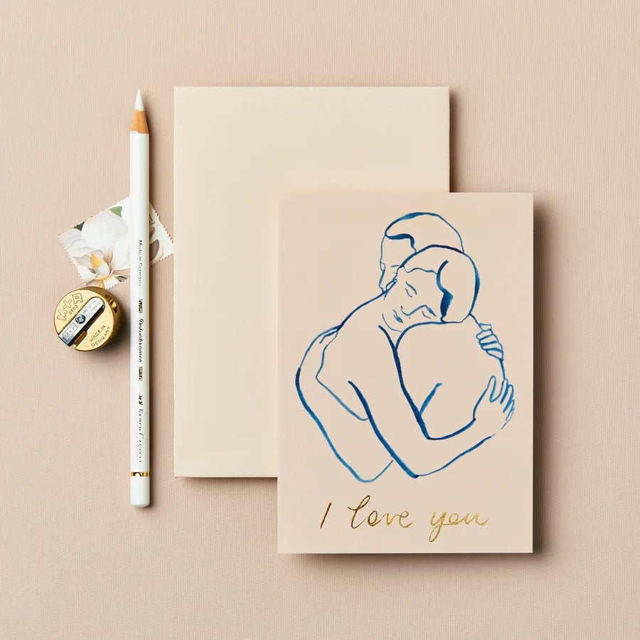 Soft beige background with a romantic greeting card featuring a minimalist blue line art of a couple embracing, and "I love you" written in gold; accented by a white pencil, flower, and tape.
