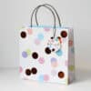 Colorful paper gift bag with polka dot pattern, handles, and matching tag, perfect for gift wrapping and celebrations.