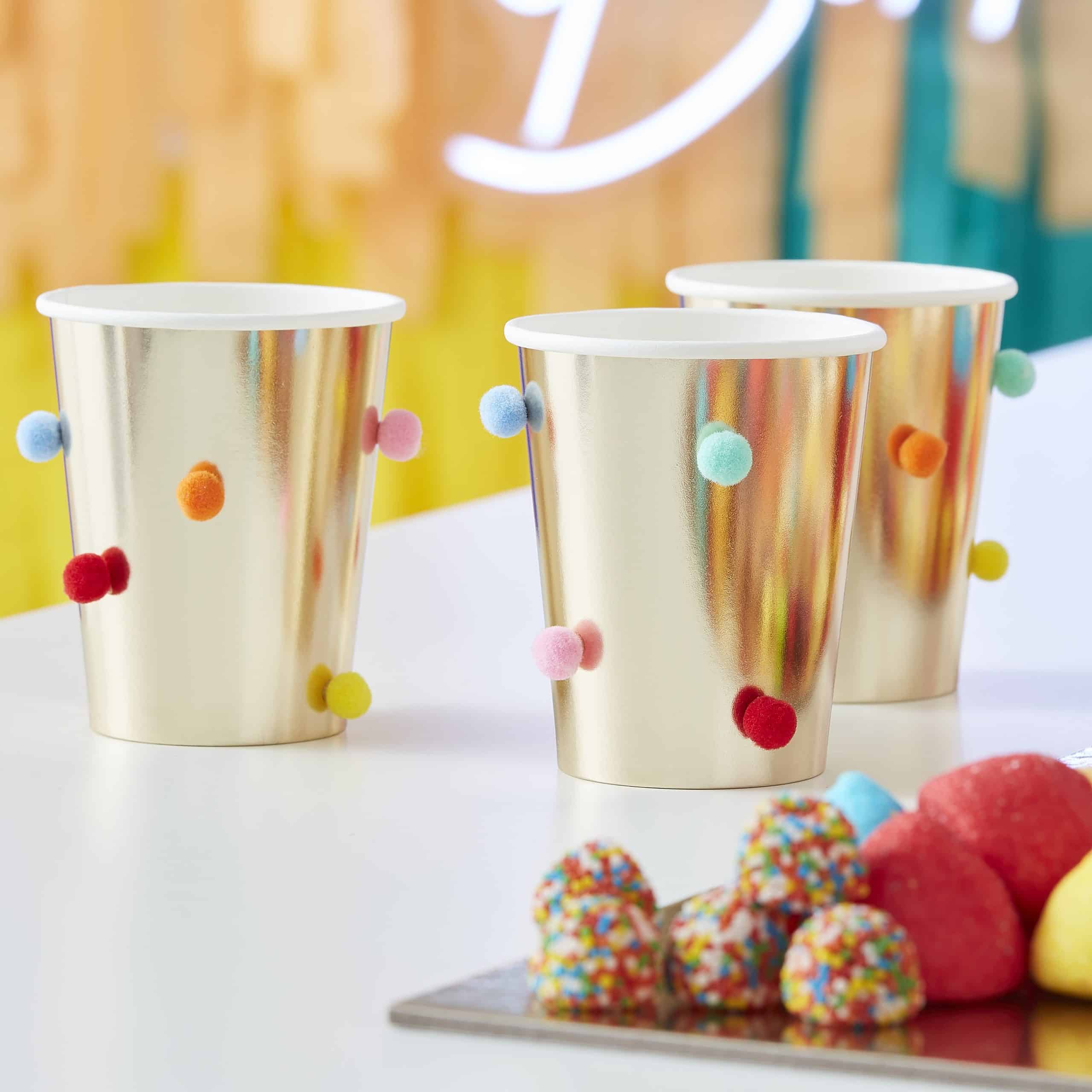 Gold metallic cups with colorful pom-pom decorations for party drinks.
