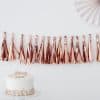 Rose gold metallic fringe garland for birthday party decor at The Wraperie. Elegant celebration adornment with festive cake and stylish accessories. Perfect for creating a fun party atmosphere.