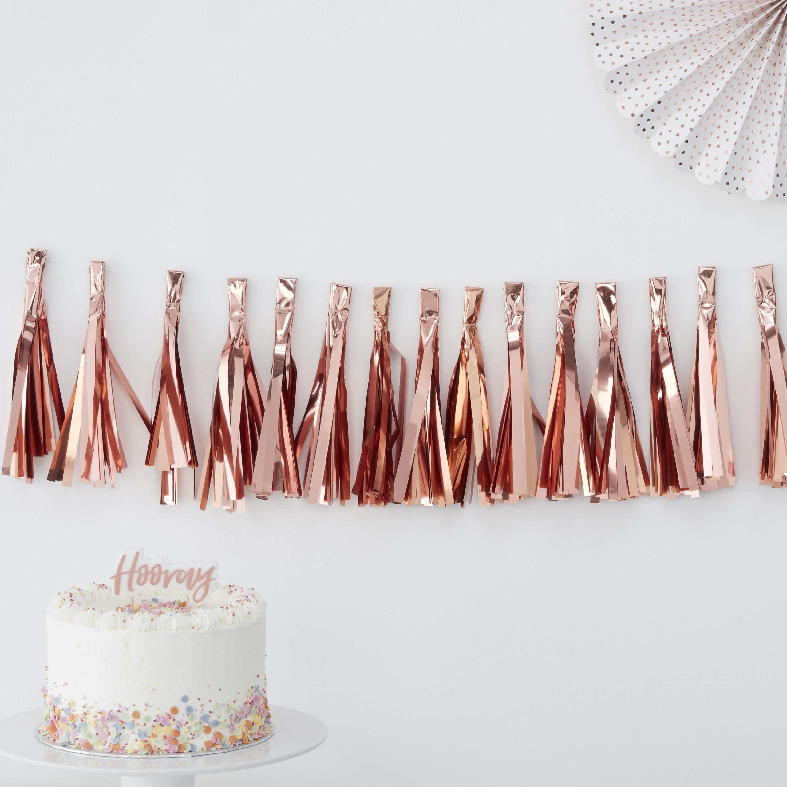 Rose gold metallic fringe garland for birthday party decor at The Wraperie. Elegant celebration adornment with festive cake and stylish accessories. Perfect for creating a fun party atmosphere.