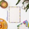 Bright colorful stationery including notepad, pen, washi tapes, paper clips, and art supplies on a white desk, promoting creative note-taking and organization with The Wraperie.