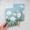 Cosmetic or stationery notebooks with floral design, featuring white flowers and green foliage on pastel blue backgrounds, perfect for creative journaling or gift giving.