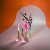 Elegant pink and white candles with a flower arrangement in a textured glass vase, set against a vibrant orange and soft pink background for a modern decorative display.