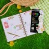 Colorful planner and sticker sheets for organization and fun, set on vibrant grass with decorative flowers and picnic accessories. Perfect for cheerful planning and creative activities.