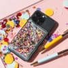 Colorful phone case with glitter and confetti for iPhone, surrounded by crafting supplies and decorative confetti on pastel pink background. Perfect for phone accessories and DIY decor.