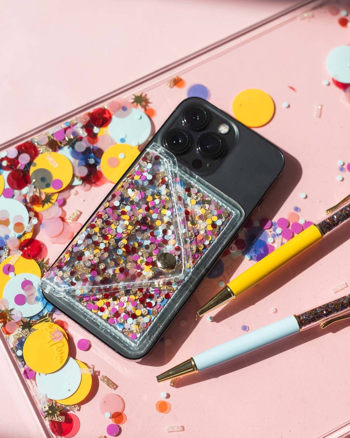 Colorful phone case with glitter and confetti for iPhone, surrounded by crafting supplies and decorative confetti on pastel pink background. Perfect for phone accessories and DIY decor.