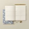Organized weekly planner with a blue floral notebook, gold pen, paper clips, and a mini stapler on a neutral background, perfect for meal planning or scheduling at The Wraperie.