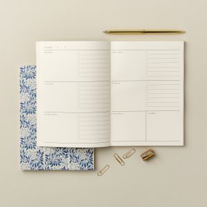 Organized weekly planner with a blue floral notebook, gold pen, paper clips, and a mini stapler on a neutral background, perfect for meal planning or scheduling at The Wraperie.