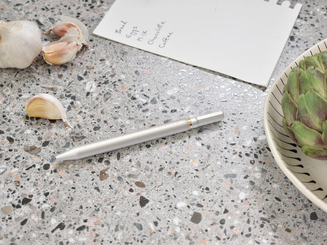 Fresh garlic cloves and a handwritten recipe note on a speckled countertop, with an artichoke in a decorative bowl, highlighting the fresh ingredients and culinary focus of The Wraperie.