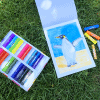 Colorful pastel chalks and a watercolor painting of a penguin on paper displayed on green grass.