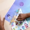 Colorful space-themed sticker set being applied to a transparent surface at The Wraperie, showcasing fun space illustrations for creative wrapping and decorating ideas.