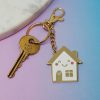 Shiny gold house-shaped keychain with a cute smiling face, pink cheeks, and a heart details, attached to a key and a chain, on pastel pink and blue background, ideal for home or keys decoration.