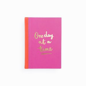 Pink notebook with gold "One day at a time" quote for motivation and positivity.
