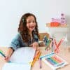 Bright young girl with curly hair coloring at a desk with colorful markers and paper, enjoying arts and crafts activities.
