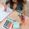 Colorful markers and stationery on wooden table with a young girl drawing rainbow, promoting creative craft supplies at The Wraperie.