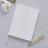 Elegant white wedding planner notebook with embossed lettering, accompanied by a gold pen and white orchids for wedding planning preparations.