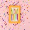 Bright pastel-colored ink pens set included in a cheerful orange box with white polka dots, surrounded by colorful confetti on a pink background, ideal for creative writing or journaling.