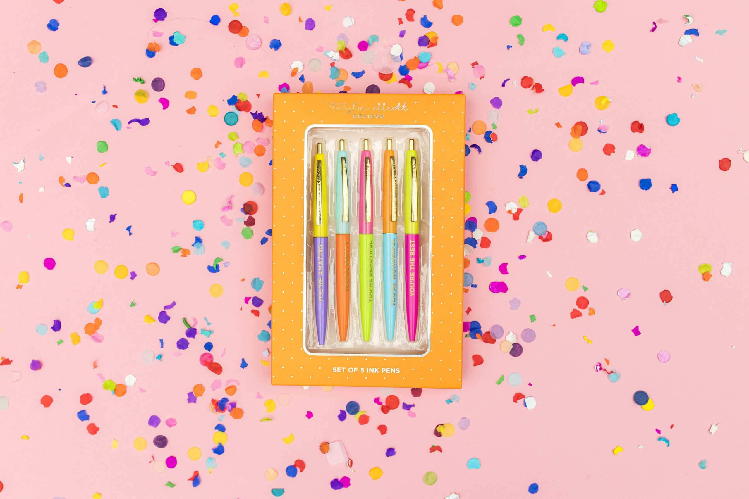 Bright pastel-colored ink pens set included in a cheerful orange box with white polka dots, surrounded by colorful confetti on a pink background, ideal for creative writing or journaling.