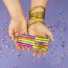 Colorful pens with motivational phrases held by a person with gold bangles on a purple background with scattered confetti.