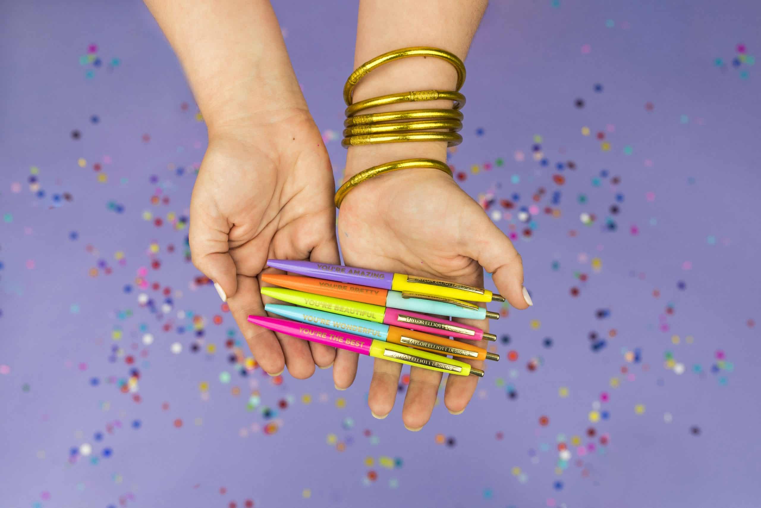 Colorful pens with motivational phrases held by a person with gold bangles on a purple background with scattered confetti.
