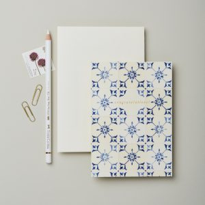 Elegant congratulation card with blue floral pattern, accompanied by a white envelope, gold paperclips, a decorative stamp, and a textured white pencil on a neutral background. Perfect for special occasions and celebrations.