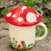 Bright ceramic mug designed to look like a whimsical mushroom with a red cap and white spots, set outdoors among greenery and soil.