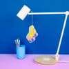 Colorful paper shoe craft hanging from a white desk lamp on a vibrant blue background, representing creative DIY projects at The Wraperie.