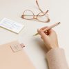 Glasses with rose gold frames, handwritten notes, pink notebook, and pen on a clean desk, showcasing stylish organization and office essentials at The Wraperie.