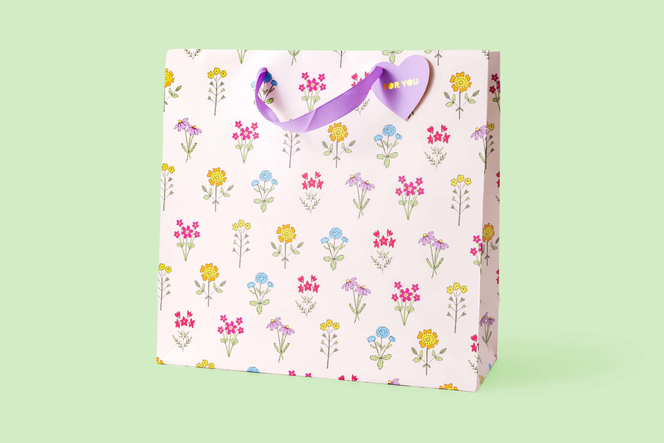 Pink floral gift bag with purple ribbon handle, decorated with colorful botanical illustrations, perfect for special occasions, gift-giving, and celebrations at The Wraperie.