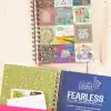 Colorful motivational quote notebooks with floral and whimsical designs, promoting mental health and positivity, displayed on a white wooden background.