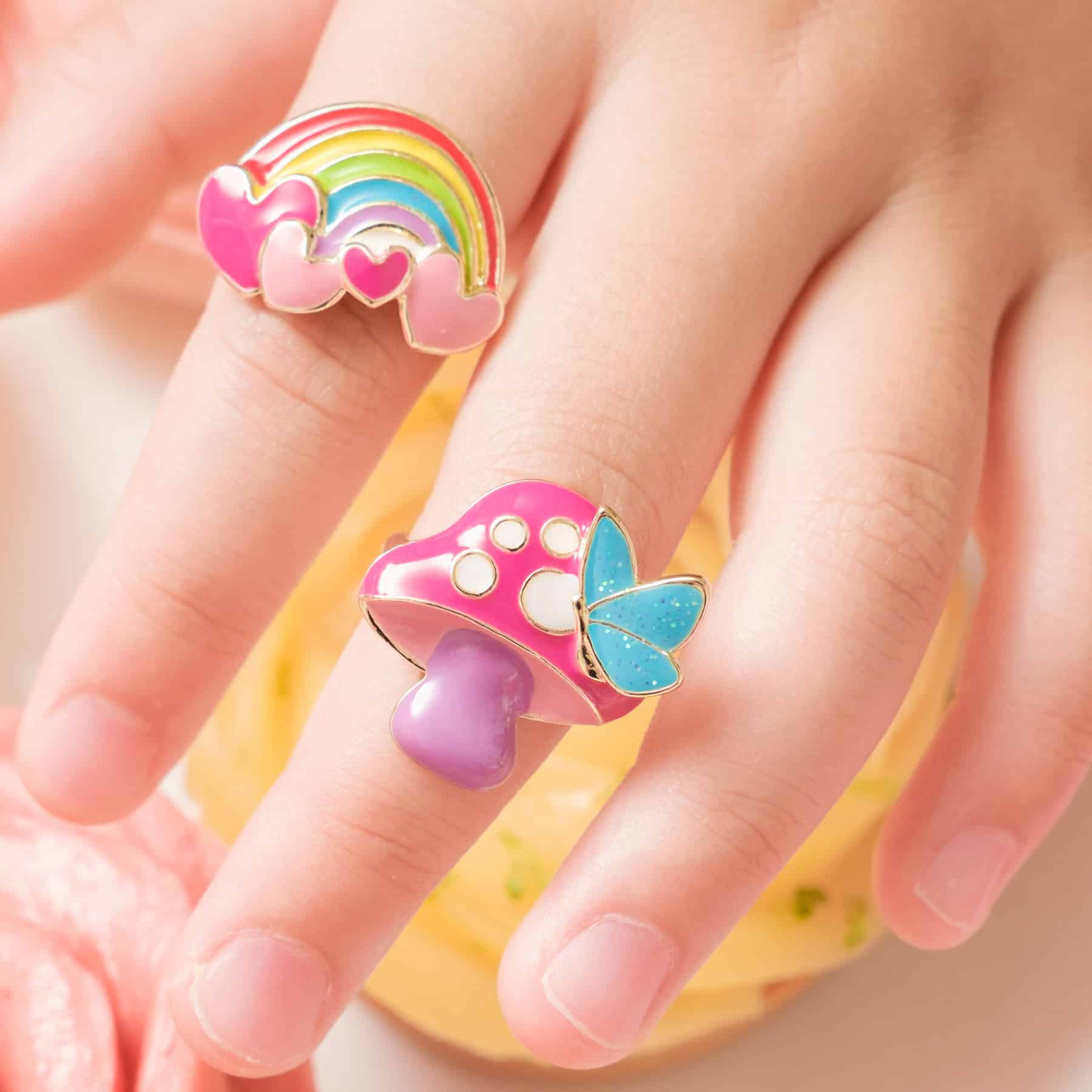 Colorful rainbow and mushroom enamel rings with butterfly accents on a hand, showcasing fun, whimsical jewelry available at The Wraperie. Perfect accessories for playful, vibrant style.
